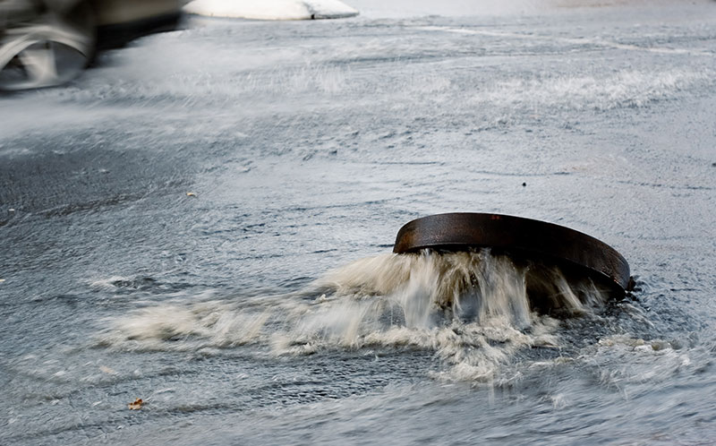 Foto von einem Kanalschacht, aus dem bei starken Regenfall Wasser auf die Straße heraussprudelt