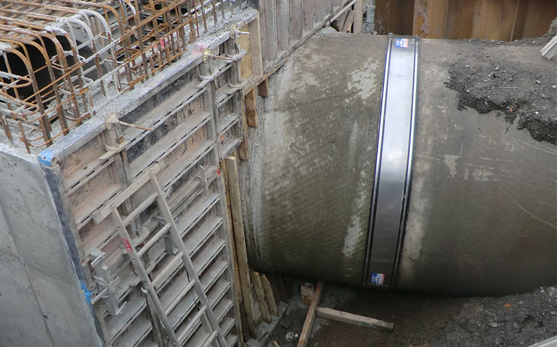 Foto von einer Tiefbau-Baustelle. An einem Betonrohr mit mehreren Metern Durchmesser wurde eine große Manschettendichtung eingebaut. Das Rohr führt in eine neue Konstruktion hinein.