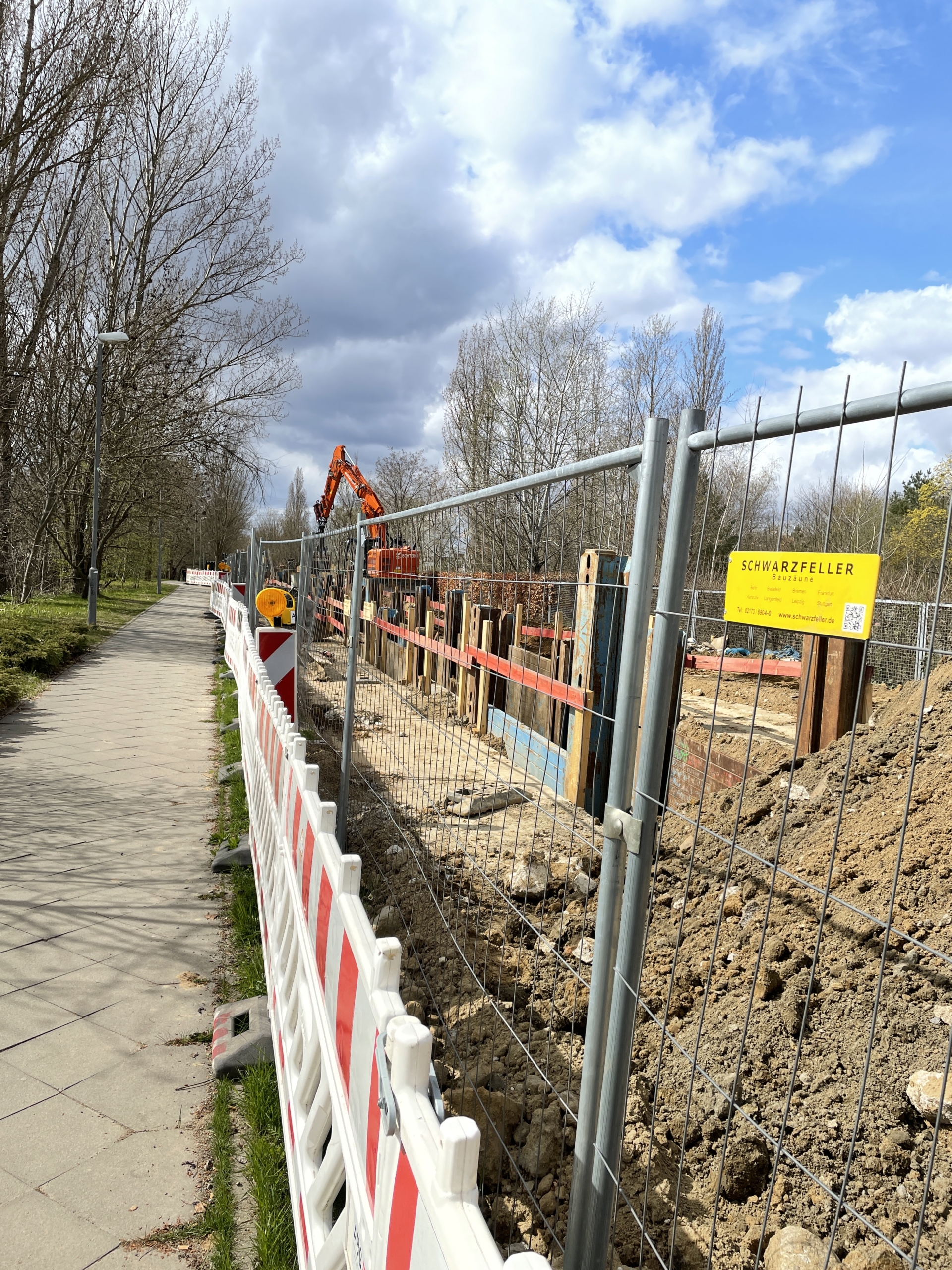 Foto einer Baustelle an einem Gehweg entlang. Bauzäune grenzen den Gehweg von einem ausgebaggerten Kanalschacht ab.